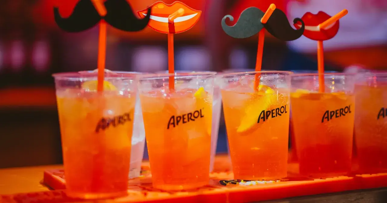 Idealne drinki na aperitif: orzeźwiające Aperole z lodem i plasterkiem pomarańczy, ozdobione zabawnymi wąsami i ustami.