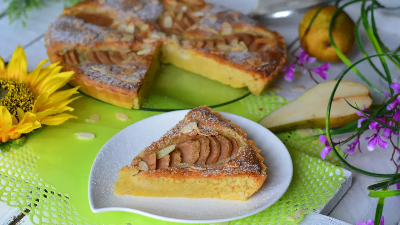Pyszna tarta z gruszkami, posypana cukrem pudrem i płatkami migdałów, prezentuje się apetycznie na talerzyku. Obok cała tarta, gruszki i słonecznik.