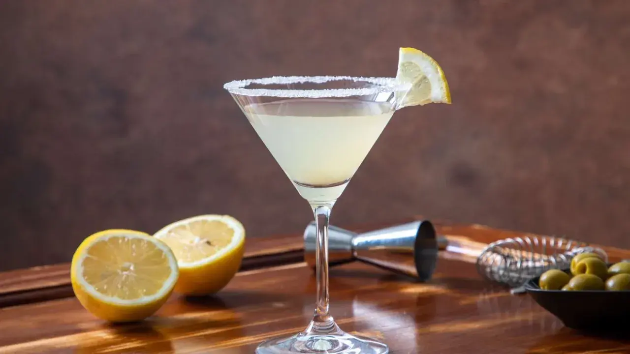 Orzeźwiający drink z martini, z solnym brzegiem kieliszka i plasterkiem cytryny. Obok leżą połówki cytryny i oliwki.