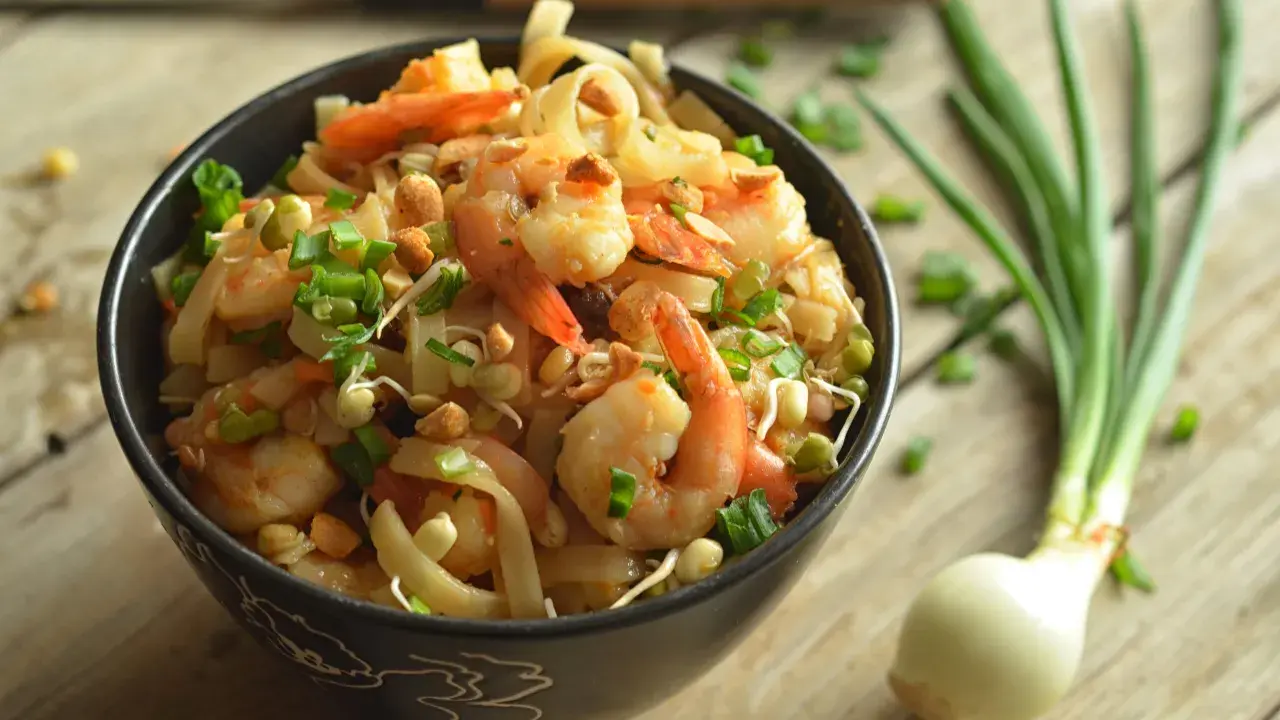 A miseczce znajduje się pyszny pad thai z krewetkami, posypany szczypiorkiem i orzeszkami. Obok leży cebulka dymka.