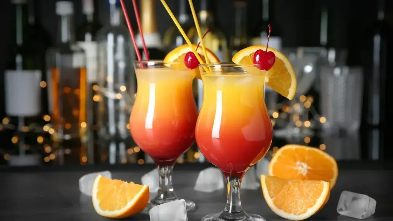 Dwa drinki z pomarańczą i wiśnią, przypominające Tequilę Sunrise. W tle bar z butelkami i światłami.