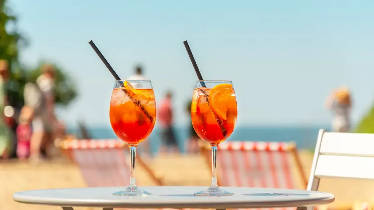 Dwa orzeźwiające aperol z tonikiem na stoliku plażowym, w tle morze i ludzie.