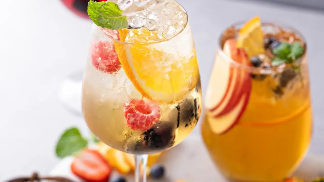 Orzeźwiający drink z prosecco, malinami, pomarańczą i miętą. Obok drugi napój z brzoskwinią i pomarańczą.