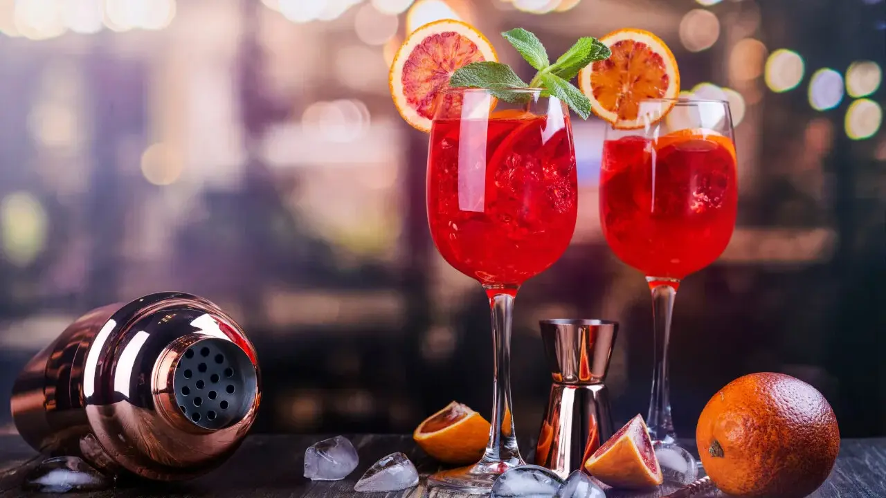 Dwa orzeźwiające drinki z białym winem, ozdobione plasterkami pomarańczy i miętą, stoją obok shakera i owoców.
