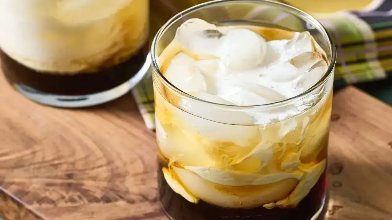 Dwa drinki White Russian z lodem i śmietanką, podane na drewnianej desce.