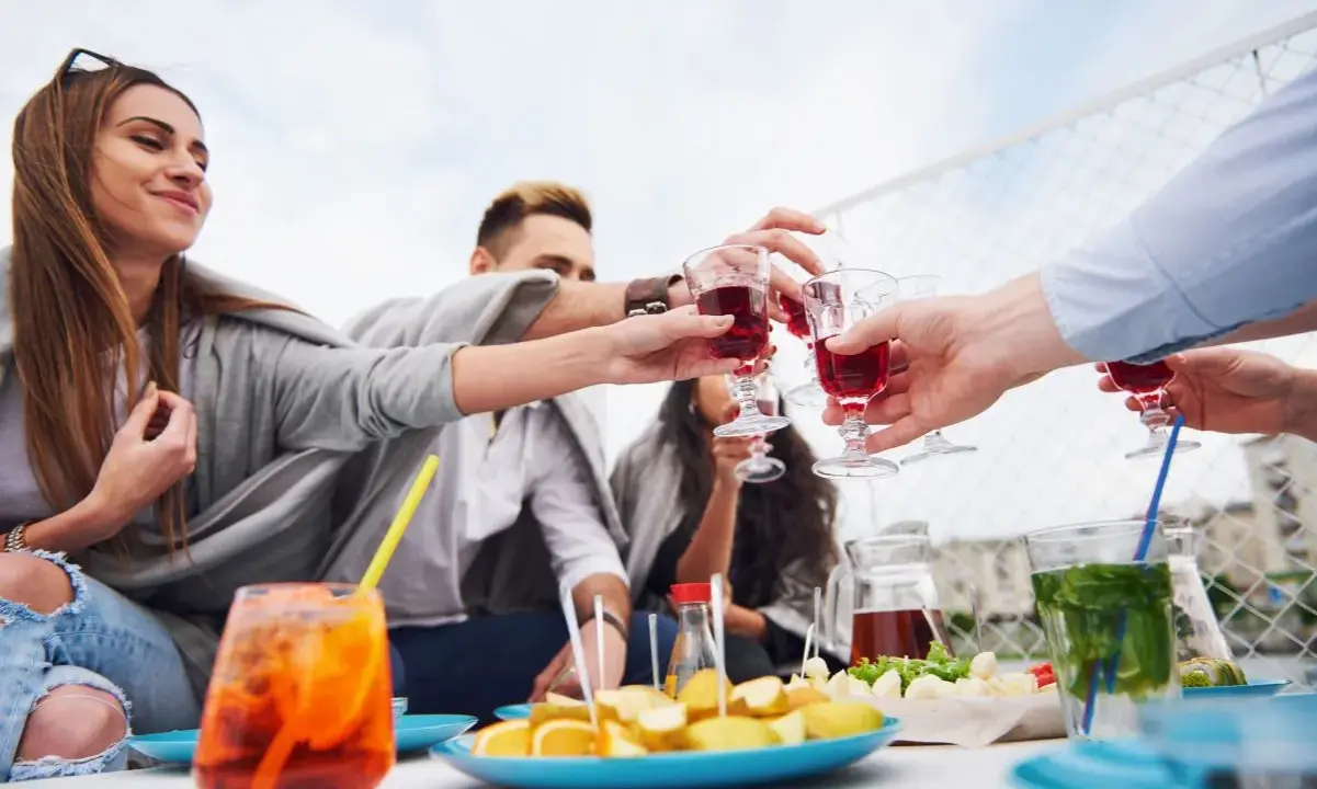 Grupa przyjaciół wznosi toast czerwonym aperitif drinkiem. Na stole owoce i inne przekąski.