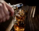 Ręka nalewa bursztynowy płyn, prawdopodobnie bourbon, do szklanek.