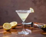 Orzeźwiający drink z martini, z solnym brzegiem kieliszka i plasterkiem cytryny. Obok leżą połówki cytryny i oliwki.