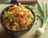 A miseczce znajduje się pyszny pad thai z krewetkami, posypany szczypiorkiem i orzeszkami. Obok leży cebulka dymka.