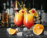 Dwa drinki z pomarańczą i wiśnią, przypominające Tequilę Sunrise. W tle bar z butelkami i światłami.