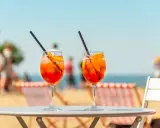 Dwa orzeźwiające aperol z tonikiem na stoliku plażowym, w tle morze i ludzie.