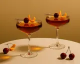 Dwa eleganckie drinki ze spirytusem, ozdobione wiśniami i skórką pomarańczową, stoją na białym blacie.