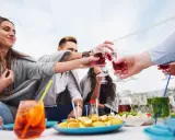 Grupa przyjaciół wznosi toast czerwonym aperitif drinkiem. Na stole owoce i inne przekąski.