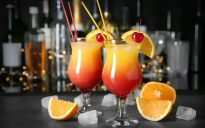 Dwa drinki z pomarańczą i wiśnią, przypominające Tequilę Sunrise. W tle bar z butelkami i światłami.