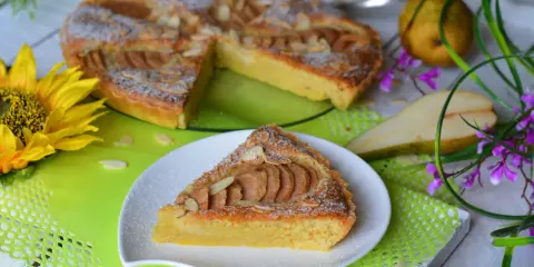Pyszna tarta z gruszkami, posypana cukrem pudrem i płatkami migdałów, prezentuje się apetycznie na talerzyku. Obok cała tarta, gruszki i słonecznik.