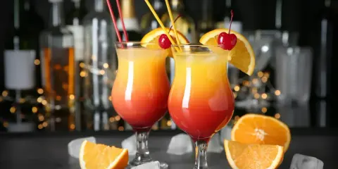 Dwa drinki z pomarańczą i wiśnią, przypominające Tequilę Sunrise. W tle bar z butelkami i światłami.