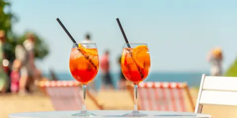 Dwa orzeźwiające aperol z tonikiem na stoliku plażowym, w tle morze i ludzie.