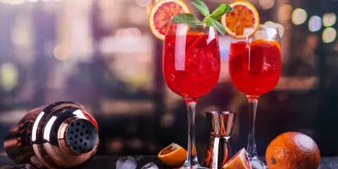 Dwa orzeźwiające drinki z białym winem, ozdobione plasterkami pomarańczy i miętą, stoją obok shakera i owoców.