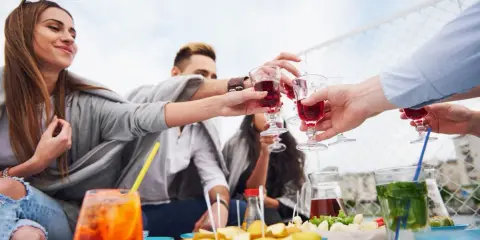Grupa przyjaciół wznosi toast czerwonym aperitif drinkiem. Na stole owoce i inne przekąski.
