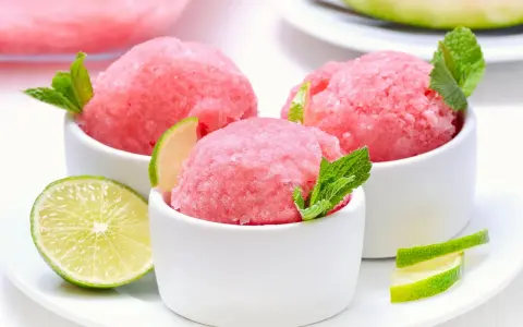 Orzeźwiający sorbet z arbuza w białych miseczkach, udekorowany listkami mięty i plasterkami limonki. Idealny na upalne dni.