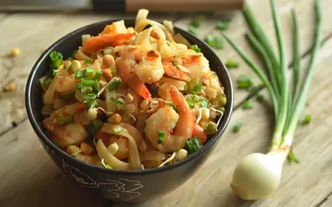 A miseczce znajduje się pyszny pad thai z krewetkami, posypany szczypiorkiem i orzeszkami. Obok leży cebulka dymka.