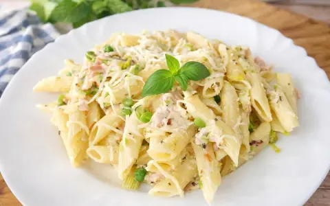 Penne z porem, groszkiem i szynką w kremowym sosie, posypane startym serem i udekorowane listkiem bazylii.