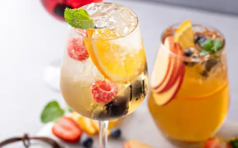 Orzeźwiający drink z prosecco, malinami, pomarańczą i miętą. Obok drugi napój z brzoskwinią i pomarańczą.