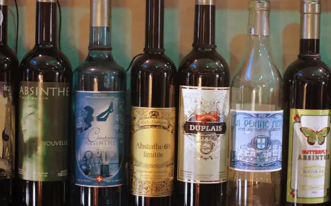 Rząd butelek z napisem "Absynt co to". Widać różne marki, m.in. Clandestine, Eichelberger, Duplais, La Bonne Mere, Butterfly Absinthe.