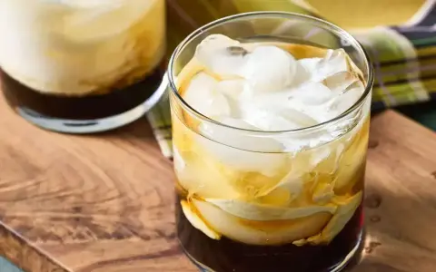 Dwa drinki White Russian z lodem i śmietanką, podane na drewnianej desce.