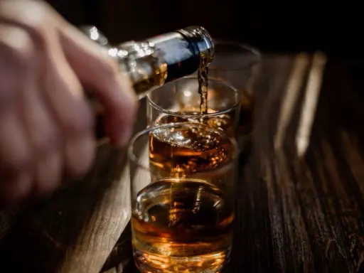 Ręka nalewa bursztynowy płyn, prawdopodobnie bourbon, do szklanek.