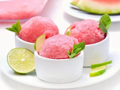 Orzeźwiający sorbet z arbuza w białych miseczkach, udekorowany listkami mięty i plasterkami limonki. Idealny na upalne dni.