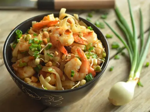 A miseczce znajduje się pyszny pad thai z krewetkami, posypany szczypiorkiem i orzeszkami. Obok leży cebulka dymka.