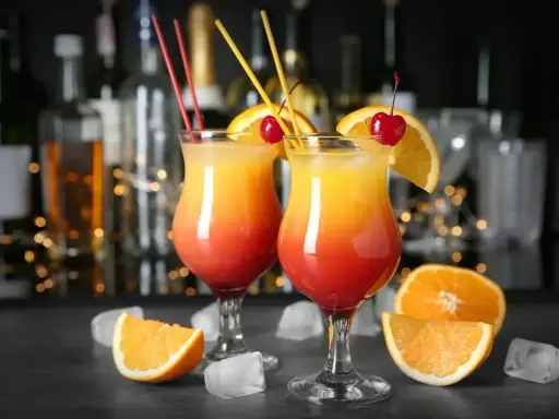 Dwa drinki z pomarańczą i wiśnią, przypominające Tequilę Sunrise. W tle bar z butelkami i światłami.