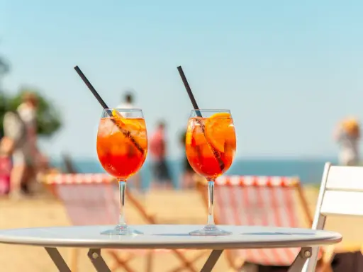 Dwa orzeźwiające aperol z tonikiem na stoliku plażowym, w tle morze i ludzie.