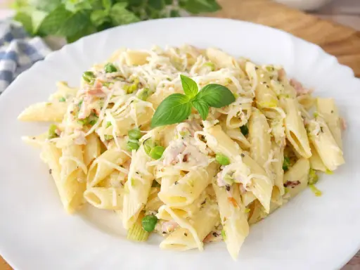 Penne z porem, groszkiem i szynką w kremowym sosie, posypane startym serem i udekorowane listkiem bazylii.