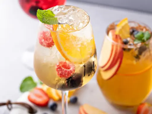 Orzeźwiający drink z prosecco, malinami, pomarańczą i miętą. Obok drugi napój z brzoskwinią i pomarańczą.