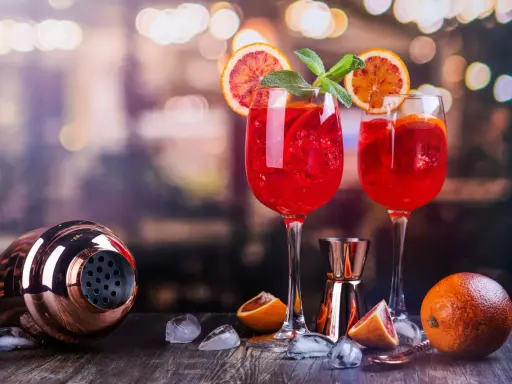 Dwa orzeźwiające drinki z białym winem, ozdobione plasterkami pomarańczy i miętą, stoją obok shakera i owoców.