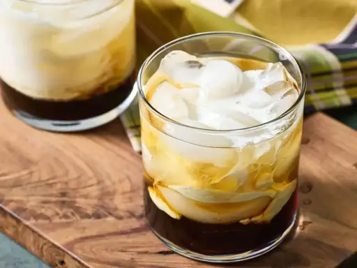 Dwa drinki White Russian z lodem i śmietanką, podane na drewnianej desce.