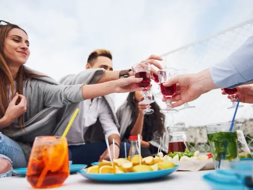 Grupa przyjaciół wznosi toast czerwonym aperitif drinkiem. Na stole owoce i inne przekąski.