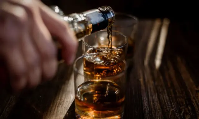 Ręka nalewa bursztynowy płyn, prawdopodobnie bourbon, do szklanek.