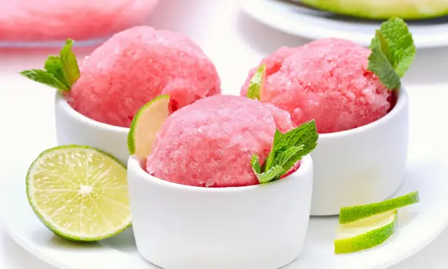 Orzeźwiający sorbet z arbuza w białych miseczkach, udekorowany listkami mięty i plasterkami limonki. Idealny na upalne dni.