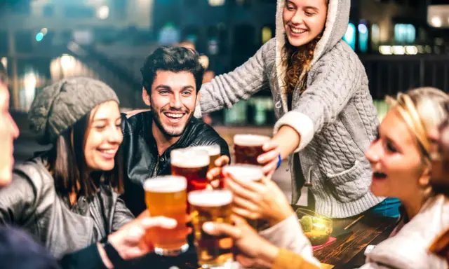 Grupa przyjaciół wznosi toasty piwem, śmiejąc się i ciesząc wieczorem. Czy piwo ma gluten? To pytanie może nurtować, ale dziś liczy się wspólna zabawa.