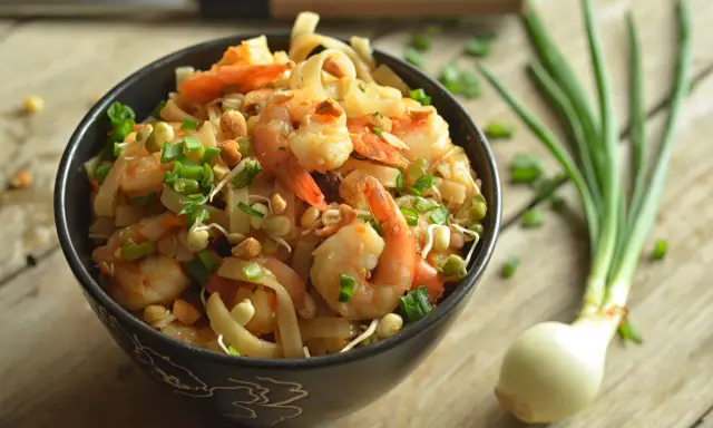 A miseczce znajduje się pyszny pad thai z krewetkami, posypany szczypiorkiem i orzeszkami. Obok leży cebulka dymka.