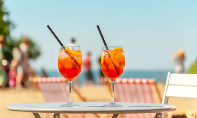 Dwa orzeźwiające aperol z tonikiem na stoliku plażowym, w tle morze i ludzie.