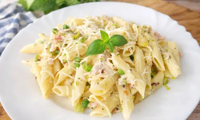 Penne z porem, groszkiem i szynką w kremowym sosie, posypane startym serem i udekorowane listkiem bazylii.