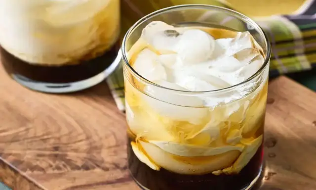 Dwa drinki White Russian z lodem i śmietanką, podane na drewnianej desce.