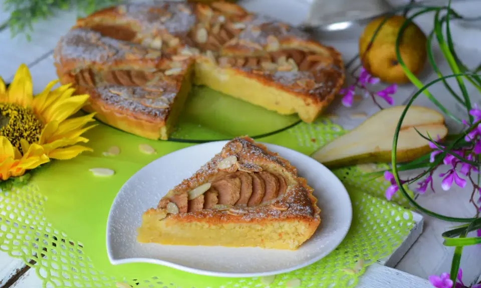 Pyszna tarta z gruszkami, posypana cukrem pudrem i płatkami migdałów, prezentuje się apetycznie na talerzyku. Obok cała tarta, gruszki i słonecznik.