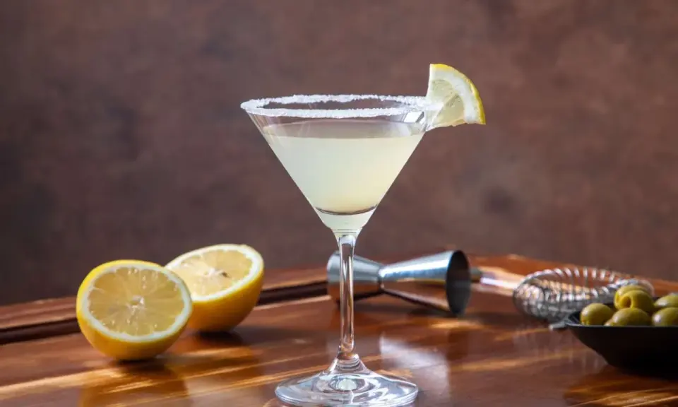 Orzeźwiający drink z martini, z solnym brzegiem kieliszka i plasterkiem cytryny. Obok leżą połówki cytryny i oliwki.