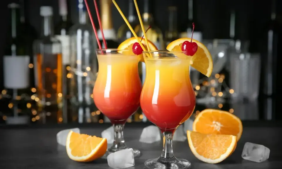 Dwa drinki z pomarańczą i wiśnią, przypominające Tequilę Sunrise. W tle bar z butelkami i światłami.