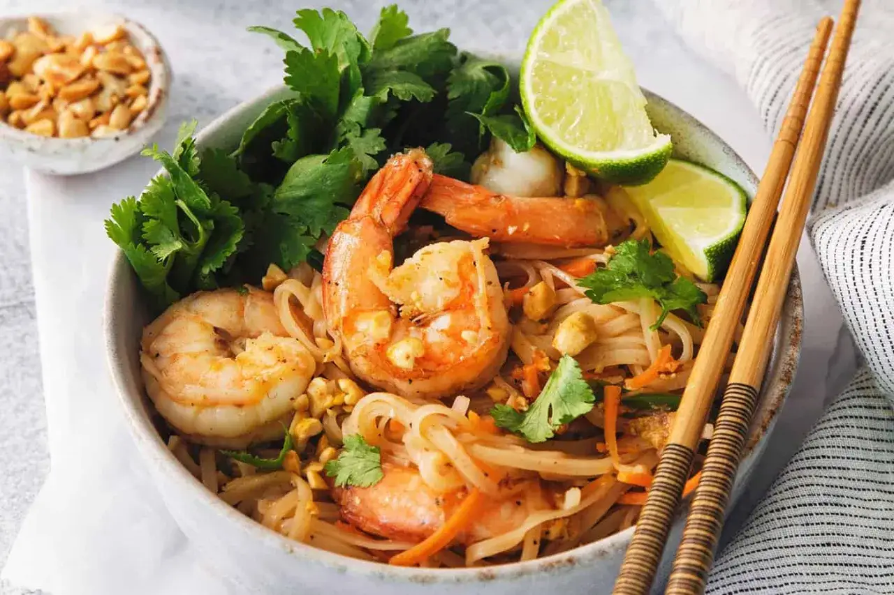 Pyszne pad thai z krewetkami, makaronem ryżowym, kolendrą i limonką. Obok miseczka z orzeszkami ziemnymi.